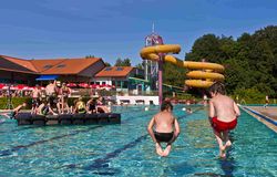 Zwei Jungen springen links vorne ins Becken vom Freibad. Rechts ist eine Gruppe junger Menschen auf eine Art Floß im Schwimmbecken zu sehen. Im Hintergrund das Gebäude des Hallenbades und eine Riesenrutsche.
