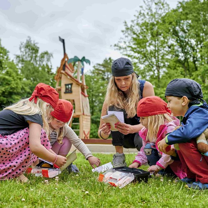 Mutter mit Kindern beim Escapegame, versammelt um die Ausrüstung auf einer Wiese 