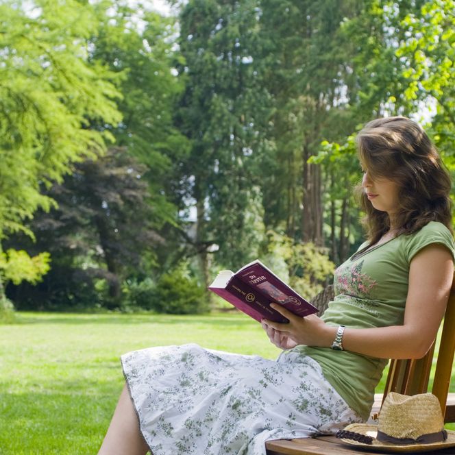 Eine junge Frau sitz lesend auf einer Parkbank. Im Hintergrund Weise und dahinter Bäume. 