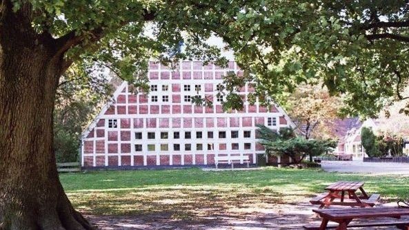 Großes Fachwerkgebäude mit weißen Fachwerk und roten Klicker von der Giebelseite aus. Im Vordergund ein großer Baum und eine Picknickbank