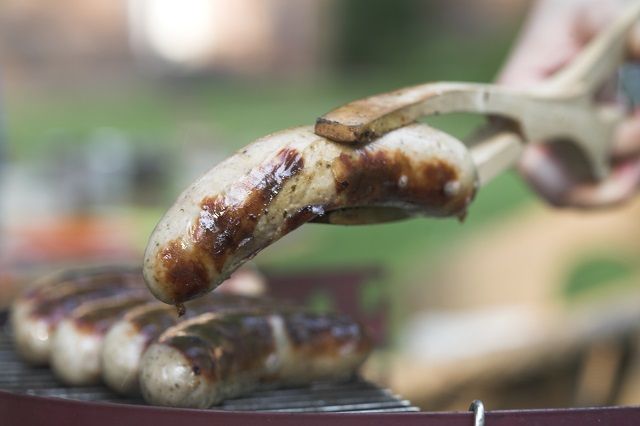 Mehrere Grillwürste auf einen Grill. Im Vordergrund eine Grillwurst in der Grillzange