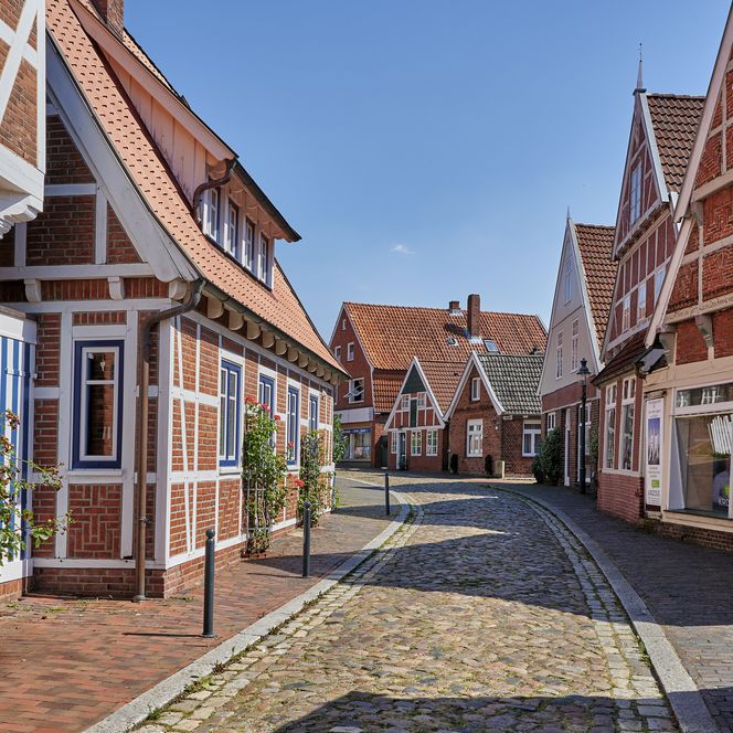 Blick in die Otterndorfer Altstadt mit Kopfsteinpflaster und rot-weißen Fachwerkhäusern unter blauem Himmel an einem Sommertag. 