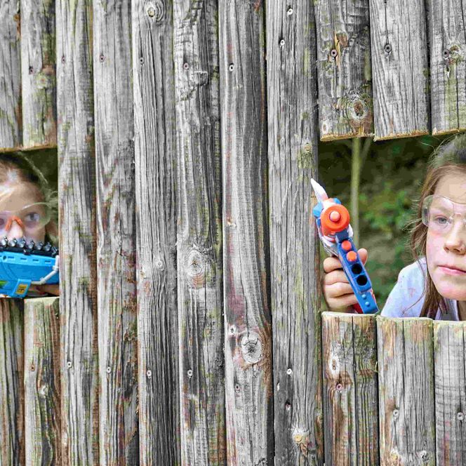 Zwei Kinder schauen durch offen Luken in einer Holzwand der Nerfarena im Spielpark WIngst