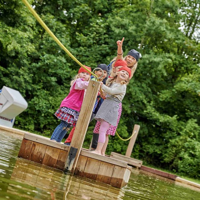 Mutter mit zwei Kindern zieht ein Spielzeugfloß mit der Leine über einen Tümpel
