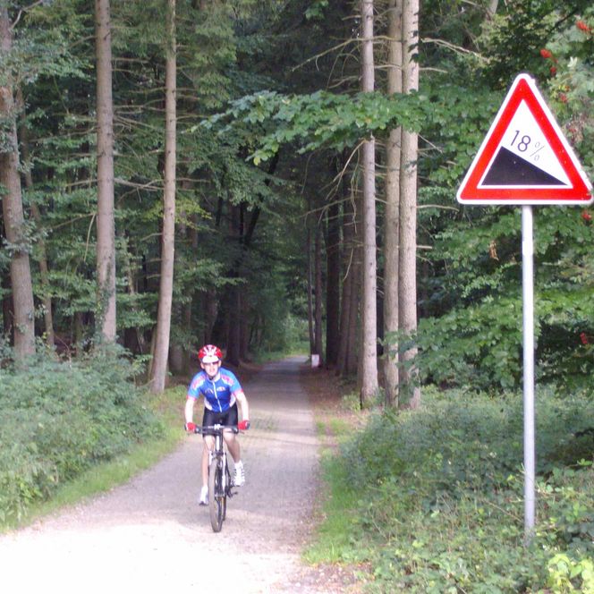 Eine MTB-Radlerin fährt einen steilen, gepflasterten Waldweg hoch, rechts ein Schild mit Steigungssatz 18%